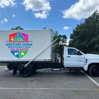 A white box truck with "Restore More Restoration" and a phone number on the side, parked in a lot with trees and a partly cloudy sky in the background.