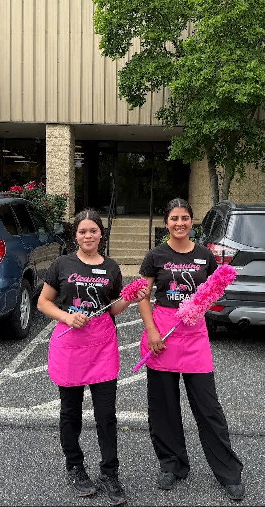 Two women wearing black shirts, pink aprons, and holding pink dusters stand in a parking lot in front of a building entrance with greenery nearby. Cars are parked beside them.