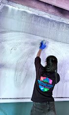 A person wearing gloves is cleaning a large, frosted glass surface with their arm raised, seen from behind. They are wearing a black shirt with colorful graphics on the back.
