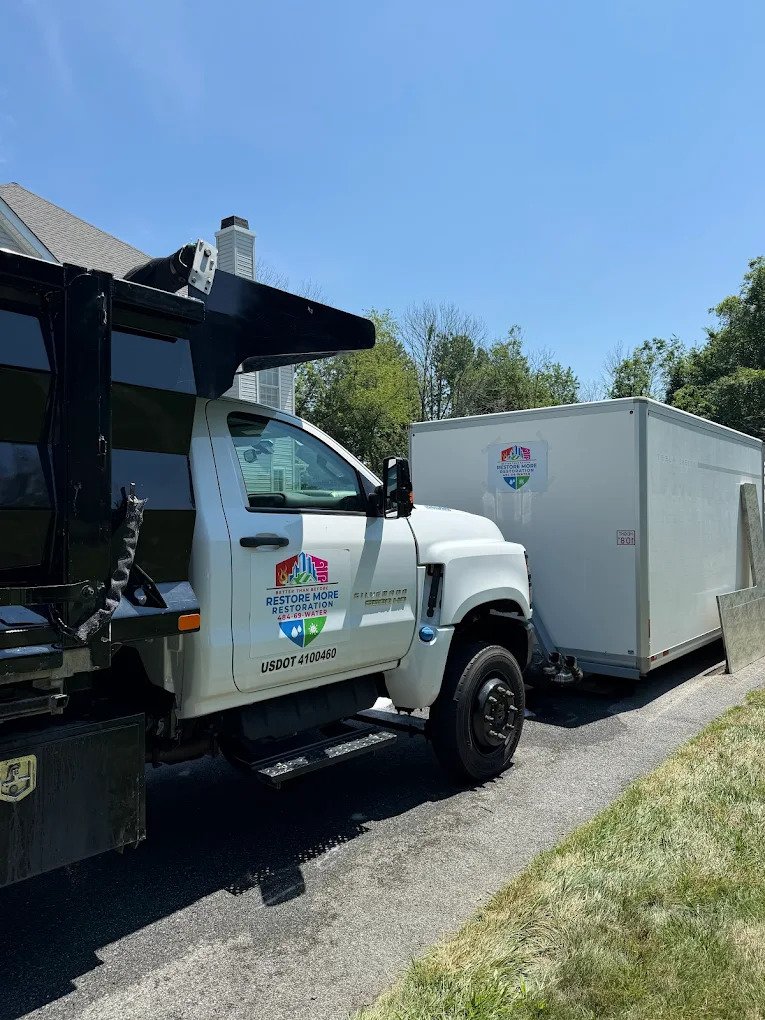 A white Restore More company truck is parked on a driveway, hitched to a white enclosed trailer. Both vehicles display the Restore More logo. Trees and a house are visible in the background on a sunny day.