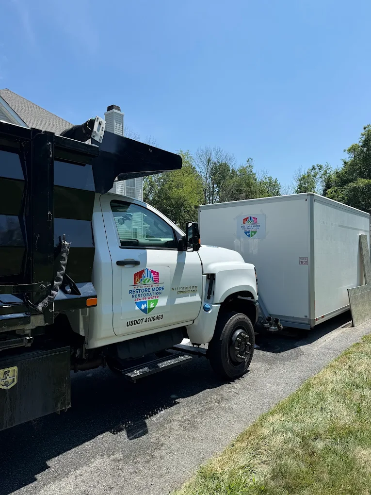 A white Restore More Restoration Services truck is parked on a driveway beside a white trailer. The sky is clear and blue, and there is a house and green trees in the background.