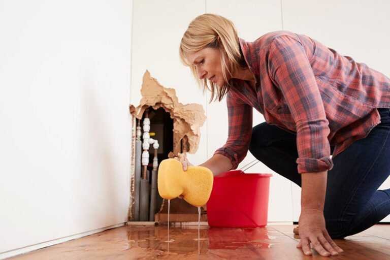 A woman in a plaid shirt and jeans uses a sponge to clean water from a wooden floor near a damaged wall. Exposed pipes are visible through the hole, indicating the need for water damage restoration. A red bucket sits beside her.