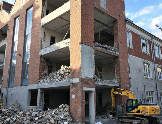 A partially demolished brick building shows signs of mold damage restoration, with exposed floors and rubble scattered on the ground. A yellow excavator is positioned next to the structure against clear blue skies in the background.