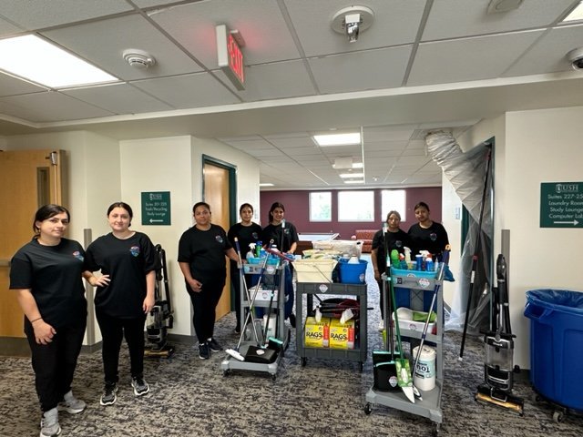 A group of six people dressed in black shirts stand in a hallway with two cleaning carts and vacuums. The hallway has white walls, carpeting, and signs indicating room locations. Cleaning supplies are visible on the carts.