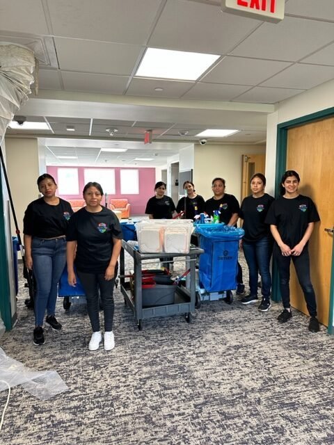 Seven women wearing matching black t-shirts stand in a hallway with cleaning carts and supplies. The setting appears to be an office or institutional building with carpeted floors and fluorescent lighting.