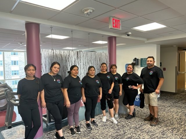 A group of eight people—seven women and one man—all wearing matching black t-shirts, stand together and smile in a brightly lit indoor area with exercise equipment and purple pillars.