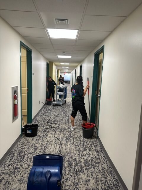 Three people are cleaning a hallway in a building. One person uses a mop near a door while others work further down the corridor. Cleaning equipment, buckets, and a floor dryer are visible along the carpeted hall.