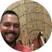 A smiling man holding a small axe stands in front of a wooden target with visible rings and marks, suggesting he has been axe throwing. A smiling man holding a small axe stands in front of a wooden target with visible rings and marks, suggesting he has been axe throwing.
