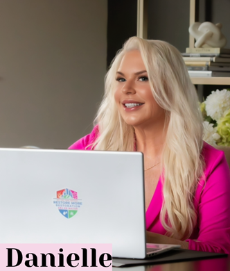 A smiling woman with long blonde hair wearing a pink blazer sits at a desk with a laptop. The laptop shows a colorful logo, and "Danielle" is in bold text. Books and flowers are in the background, hinting at her role in a top Restoration Company.