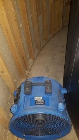 A blue industrial fan sits on a concrete floor near an unfinished wooden wall with exposed studs and insulation. The area appears to be under construction or renovation.