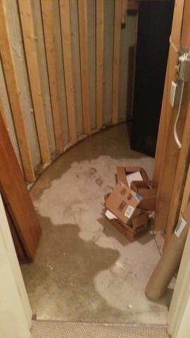 A small unfinished room with exposed wooden wall studs, a concrete floor with a damp water stain, and a pile of broken-down cardboard boxes on the right side.