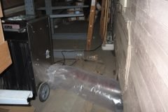 A cluttered storage area with shelves, a black cabinet on wheels, scattered plastic wrapping, and various items on the floor, viewed from a narrow aisle beside a wooden wall.