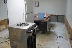 A room with a concrete floor, exposed drywall along the bottom of the walls, a refrigerator, a metal appliance, and a desk with a blue bin. The setting appears under construction or damaged.